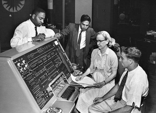 A imagem de 1960 mostra quatro pessoas, 3 homens e uma mulher, sentados em torno de um antigo console de computador conhecido como UNIVAC 1. A mulher se chama Grace Hopper e segura um conjunto de folhas de papel com dados técnicos.