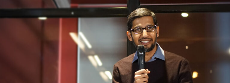 A imagem apresenta Sundar Pichai, CEO do Google, sorrindo e olhando para a frente, segurando um microfone. Ele usa óculos e veste uma camisa azul clara com um suéter marrom.